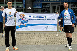 Zwei Männer mit MUKOmove-T-Shirts halten ein MUKOmove-Banner in die Kamera. Im Hintergrund sieht man den Startbereich einer Laufveranstaltung. 