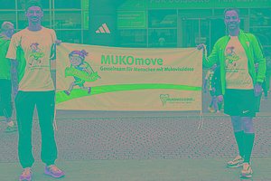 Zwei Männer mit MUKOmove-T-Shirts halten ein MUKOmove-Banner in die Kamera. Im Hintergrund sieht man den Startbereich einer Laufveranstaltung. 