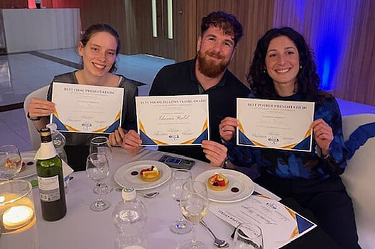Gruppenbild an einem noch gedeckten Tisch, zwei Frauen und ein Mann halten Urkunden in die Kamera. 