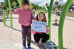 Zwei Mädchen, unter zehn Jahre alt, halten ein Schild ("Mukoviszidose braucht mehr Aufmerksamkeit") auf dem Spielplatz in die Kamera. 
