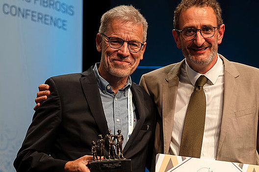 Stephan Kruip erhält den Patient Advocacy Award von CF Europe Zwei ältere Männer auf einer Bühne, einer hält einen Preis in der Hand.