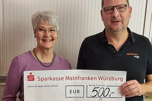 Eine ältere Frau und ein Mann halten einen symbolischen Spendenscheck über 500 Euro in die Kamera. 
