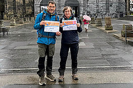 Mann und Frau halten in einer Stadt und im Regen ihre MUKOmove-Startnummern in die Kamera