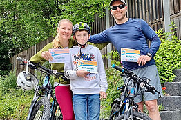 Eine Frau, ein Mann und ein Junge (vielleicht zwölf Jahre alt) stehen neben Fahrrädern im Wald und halten MUKOmove-Startnummern in der Hand. Der Junge trägt einen Fahrradhelm. 