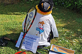 Ein Stoffpinguin trägt eine MUKOmove-T-Shirt, an ihm lehnt ein Tennisschläger. 