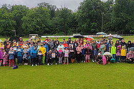 Eine große Gruppe von Menschen, die teils Regenschirme und Regenjacken tragen, weil das Wetter sichtbar schlecht ist, stehen auf einer Wiese. Einige von ihnen halten MUKOmove-Startnummern in der Hand. 