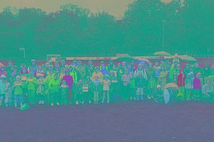 Eine große Gruppe von Menschen, die teils Regenschirme und Regenjacken tragen, weil das Wetter sichtbar schlecht ist, stehen auf einer Wiese. Einige von ihnen halten MUKOmove-Startnummern in der Hand. 