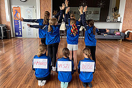 In einem Tanzstudio stehen mehrere Mädchen mit dem Rücken zur Kamera. Sie tragen blaue Shirts und auf ihren Rücken kleben Zettel, auf denen Mukoviszidose zu lesen ist. 