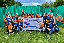 Eine Gruppe von etwa 35 Menschen steht vor einer grünen Plane, daneben sieht man Zielscheiben. Einige Menschen halten Bogen in der Hand. Außerdem halten sie ein Banner mit der Aufschrift MUKOmove in die Kamera. Die Menschen tragen blaue oder orangene T-Shirts. 