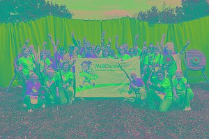 Eine Gruppe von etwa 35 Menschen steht vor einer grünen Plane, daneben sieht man Zielscheiben. Einige Menschen halten Bogen in der Hand. Außerdem halten sie ein Banner mit der Aufschrift MUKOmove in die Kamera. Die Menschen tragen blaue oder orangene T-Shirts. 