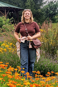 Porträt einer Frau, die in einem Park steht, man sieht viele bunte Blumen, sie trägt eine lange Hose und ein T-Shirt. 