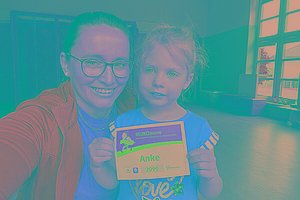 Eine Mutter (grüne Sportjacke) und ihr Kind (rotes T-Shirt) stehen in einem Raum, der nach einem Physiotherapieraum aussieht. Das Kind hält eine MUKOmove-Startnummer in der Hand, auf der "Anke" steht. 