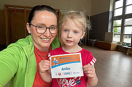 Eine Mutter (grüne Sportjacke) und ihr Kind (rotes T-Shirt) stehen in einem Raum, der nach einem Physiotherapieraum aussieht. Das Kind hält eine MUKOmove-Startnummer in der Hand, auf der "Anke" steht. 