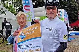 Eine Frau und ein Mann (mit Fahrradhelm und Fahrradkleidung) halten ein Schild "MUKOmove - Ich bin dabei. Du auch?" in die Kamera. Im Hintergrund sieht man, dass ein Radrennen stattfindet. 
