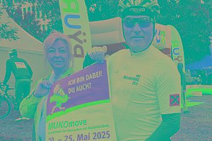Eine Frau und ein Mann (mit Fahrradhelm und Fahrradkleidung) halten ein Schild "MUKOmove - Ich bin dabei. Du auch?" in die Kamera. Im Hintergrund sieht man, dass ein Radrennen stattfindet. 