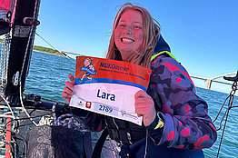 Eine Frau sitzt auf einem Segelboot, im Hintergrund sieht man Meer, blauen Himmel und eine Brücke. Die Frau hat eine Startnummer für den MUKOmove in der Hand. Darauf steht "Lara". 