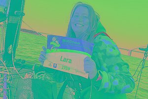Eine Frau sitzt auf einem Segelboot, im Hintergrund sieht man Meer, blauen Himmel und eine Brücke. Die Frau hat eine Startnummer für den MUKOmove in der Hand. Darauf steht "Lara". 