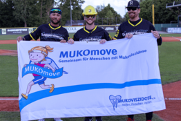 Drei Männer in Baseball-Kleidung stehen auf einem Sportplatz und halten ein MUKOmove-Banner in die Kamera. 