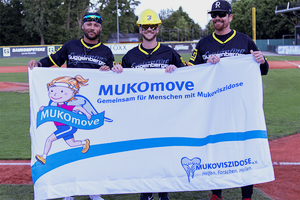 Drei Männer in Baseball-Kleidung stehen auf einem Sportplatz und halten ein MUKOmove-Banner in die Kamera. 