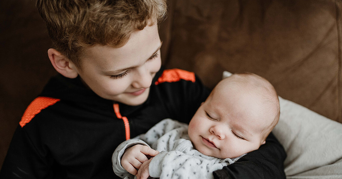 Ein achtjähriger Junge hält seinen Babybruder im Arm und schaut ihn an.