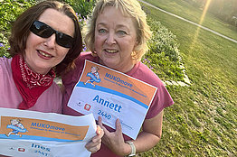 Zwei Frauen stehen auf einer Wiese oder einem Feld und halten ihre MUKOmove-Startnummern in die Kamera. 