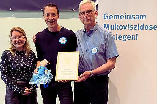 Dominik Klein ist 20 Jahre Schutzengel für Menschen mit Mukoviszidose. Eine jüngere Frau (links) und ein älterer Mann (rechts) übergeben eine Urkunde und eine Stoffpuppe mit Handball-Kleidung an einen Mann in dunkler Kleidung in der Mitte.