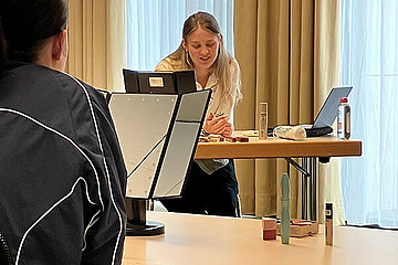 Frau auf einem Podium, auf einem Tisch stehen Schminkutensilien. 