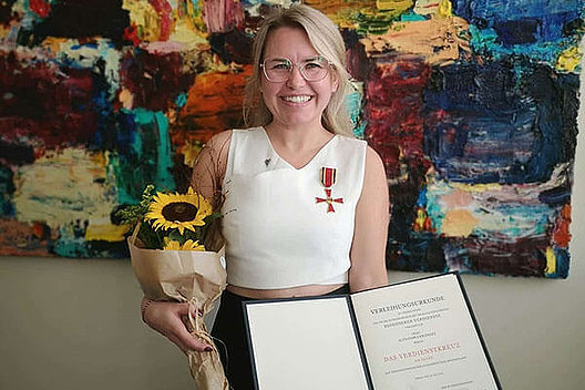 Bundesverdienstkreuz für Alexandra Kramarz Eine Frau mit weißem Oberteil hält eine Sonnenblume und eine Urkunde in die Kamera. Sie trägt einen Orden.