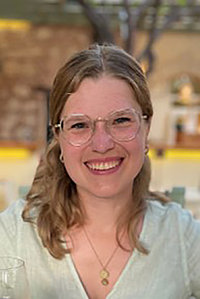 Junge blonde Frau lacht in die Kamera, sie trägt eine Brille, ihre Haare sind lang. Sie trägt ein helles Shirt. 