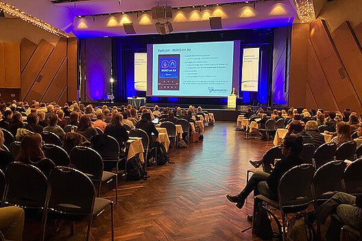 Blick ins Plenum Vortragssaal mit vielen Menschen, fotografiert von hinten. Man sieht vorne auf dem Podium jemanden stehen, der gerade einen Vortrag hält. Auf der Power-Point-Folie vorne ist "Podcast MUKO on Air" zu lesen.