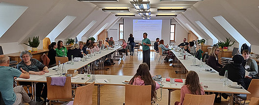 Blick in den Seminarraum Seminarraum mit Tischen in U-Form, an denen Menschen sitzen und zum Referenten nach vorne blicken.