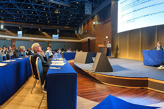 Blick von der Seite in einen Konferenzraum, Menschen im Publikum schauen auf die Bühne. 