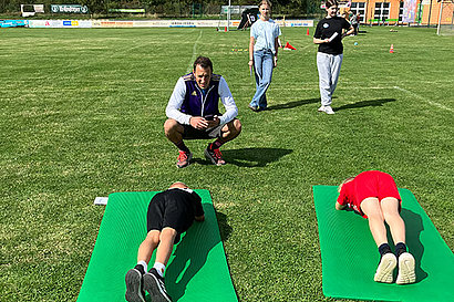 Ein Mann hockt auf einem Sportplatz vor zwei Jungen, die auf Yogamatten gerade Planks machen.