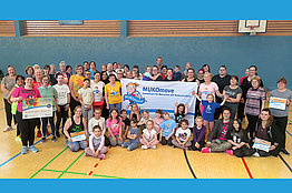 Große Gruppe Menschen (Kinder und Erwachsene) in der Turnhalle mit MUKOmove-Banner