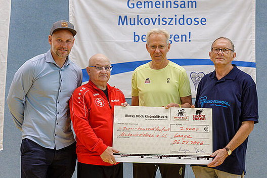 Vier Männer stehen vor einem Banner mit der Schrift "Gemeinsam Mukoviszidose besiegen" und halten einen Spendenscheck in Höhe von 2.500 Euro in die Kamera.
