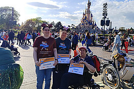 Drei Frauen stehen vor dem Schloss vom Diesneyland Paris, eine davon im Rollstuhl. Sie alle halten MUKOmove-Startnummern in die Kamera. Um sie herum sind sehr viele Menschen zu sehen. 
