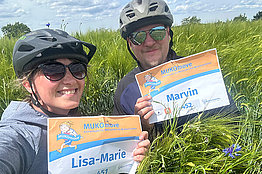 Eine Frau (sie trägt eine MUKOmove-Startnummer, auf der Lisa-Marie steht) und ein Mann (auf seiner Startnummer steht Marvin) stehen in einem Feld. Beide haben Fahrradhelme an. 