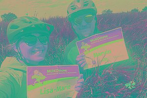 Eine Frau (sie trägt eine MUKOmove-Startnummer, auf der Lisa-Marie steht) und ein Mann (auf seiner Startnummer steht Marvin) stehen in einem Feld. Beide haben Fahrradhelme an. 