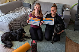 Zwei Frauen posieren in ihrem Wohnzimmer und mit ihren Hunden mit MUKOmove-Startnummern. 