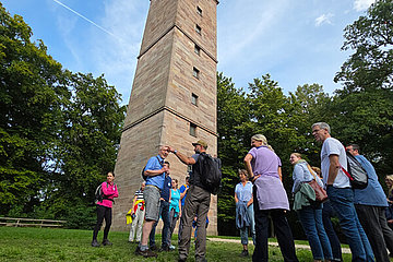 Mehrere Menschen stehen um einen gemauerten Turm herum, man sieht viele Bäume. 