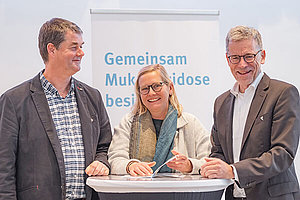 Winfried Klümpen und Miriam Schlangen aus der Geschäftsführung des Mukoviszidose e.V. mit Stephan Kruip, Vorstandsvorsitzender des Mukoviszidose e.V. (von links nach rechts), im Hintergrund sieht man die Aufschrift "Gemeinsam Mukoviszidose besiegen"