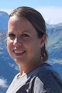 Julia Lock - Porträt. Man sieht nur ihr Gesicht, ihre etwa schulterlangen Haare sind zu einem Zopf gebunden. Im Hintergrund sieht man Berge. 