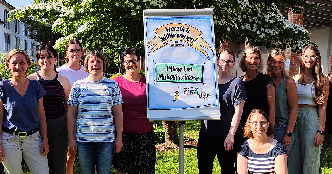 Mehrere Menschen stehen im Garten vor einem Flipchart, auf dem "Pflege bei Mukoviszidose" zu lesen ist. 
