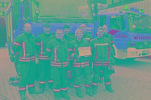 Feuerwehrleute in voller Montur halten ihre MUKOmove-Startnummer in die Kamera, im Hintergrund sieht man die Feuerwehrfahrzeuge