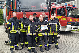 Feuerwehrleute in voller Montur halten ihre MUKOmove-Startnummer in die Kamera, im Hintergrund sieht man die Feuerwehrfahrzeuge