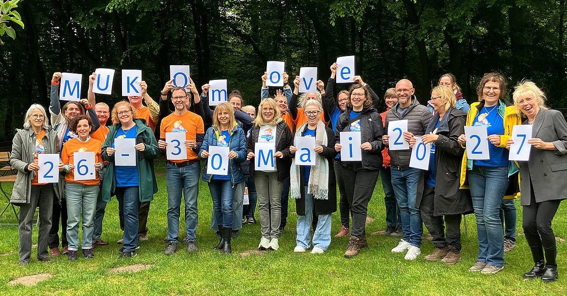 Gruppe von Menschen in MUKOmove-T-Shirts hält Schilder mit den Worten MUKOmove 26.-30. Mai 2027 in den Händen.