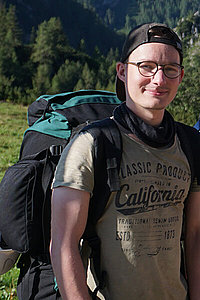 Junger Mann mit Wanderrucksack, Brille und Cappi. Er steht in der Natur. 