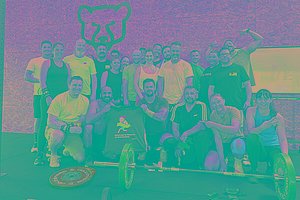Gruppe von sportlichen Menschen hat sich um Sportgeräte versammelt, im Hintergrund sieht man die Zeichnung eines Bärenkopfs. Die Menschen halten ein MUKOmove-T-Shirt in die Kamera. 