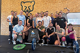 Gruppe von sportlichen Menschen hat sich um Sportgeräte versammelt, im Hintergrund sieht man die Zeichnung eines Bärenkopfs. Die Menschen halten ein MUKOmove-T-Shirt in die Kamera. 