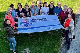 Gruppe von Frauen hält ein MUKOmove-Banner in der Hand, sie sind von oben fotografiert und stehen auf einer Wiese.